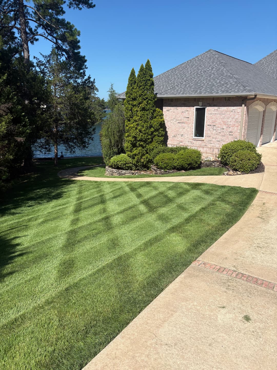 Well-manicured lawn with stripes, brick house, and lake view under clear blue sky.